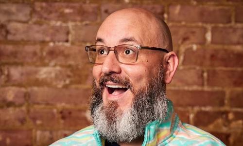 white man with very full beard and glasses smiling and looking up in front of a brick wall (picture is only from the neck up)