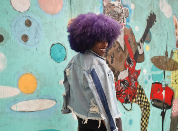 woman walking by a mural with flare-leg pants, a distressed light demin jacket, and purple afro hairstyle