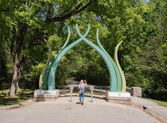 green sculptural gate entrance to old forest trail, woman walking through 