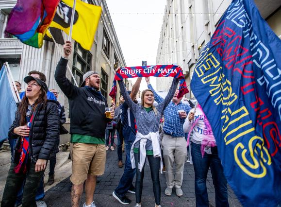 901 fc fans stand cheering in an alley during the march prior to the game. they're holding flags that say Memphis and Bluff City Mafia