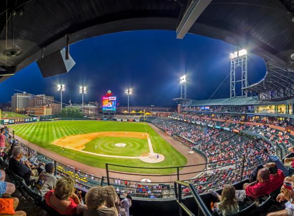 Panoramic photo of baseball stadium full of fans in the evening. The diamond is lit up by the lights