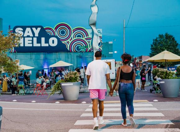 couple holding hands walks across the street in front of blue wall with say hello sign