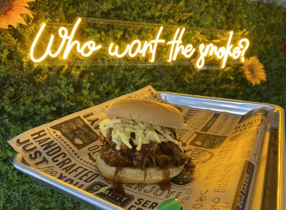 pulled pork sandwich on tray behind held up in front of "who want the smoke?" sign