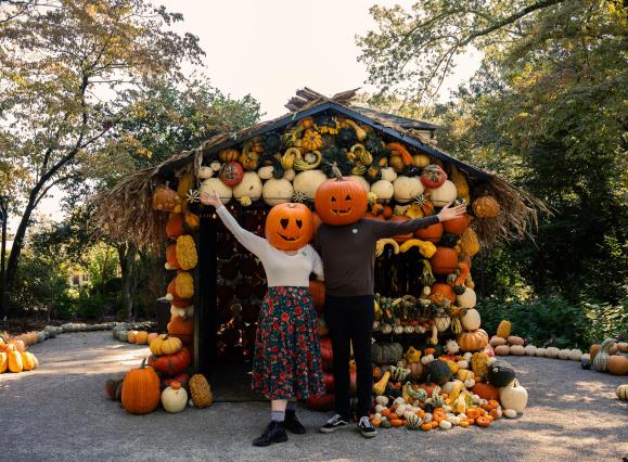 two people pose in front of a pumpkin house while wearing pumpkins on head