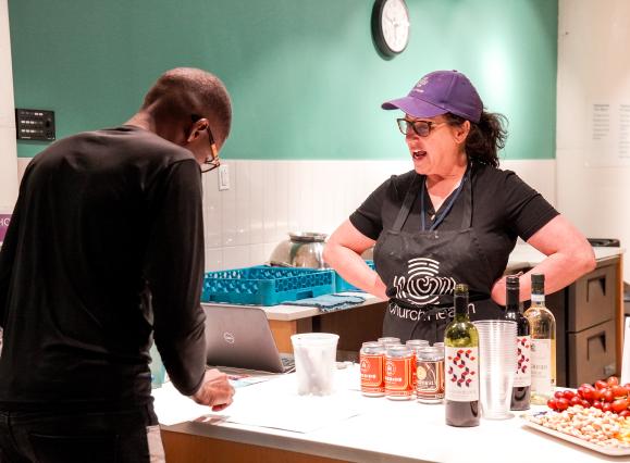 lady in purple hat standing behind table talking to man in black shirt