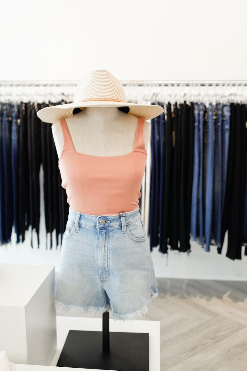 mannequin wearing pink tank and cutoff light demin jorts with a wide brim hat, rack of jeans in background