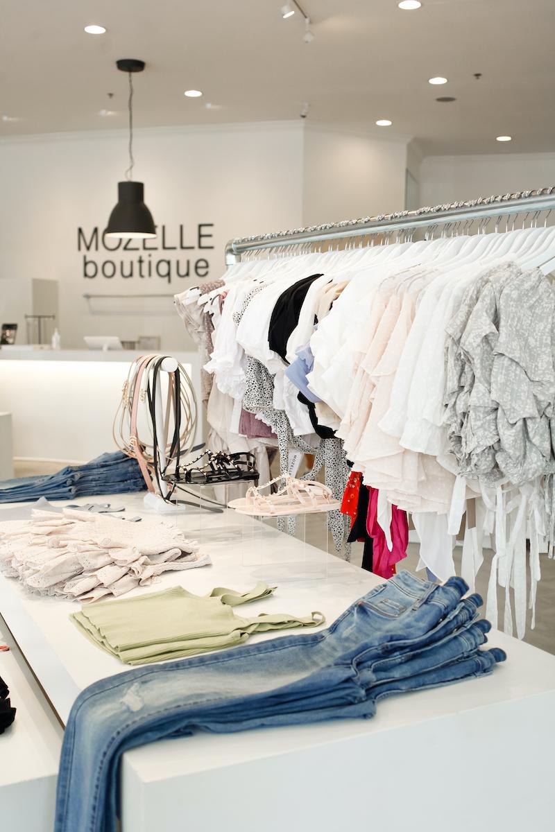light-colored summery blouses hanging on rack in bright boutique, says Mozelle on the wall