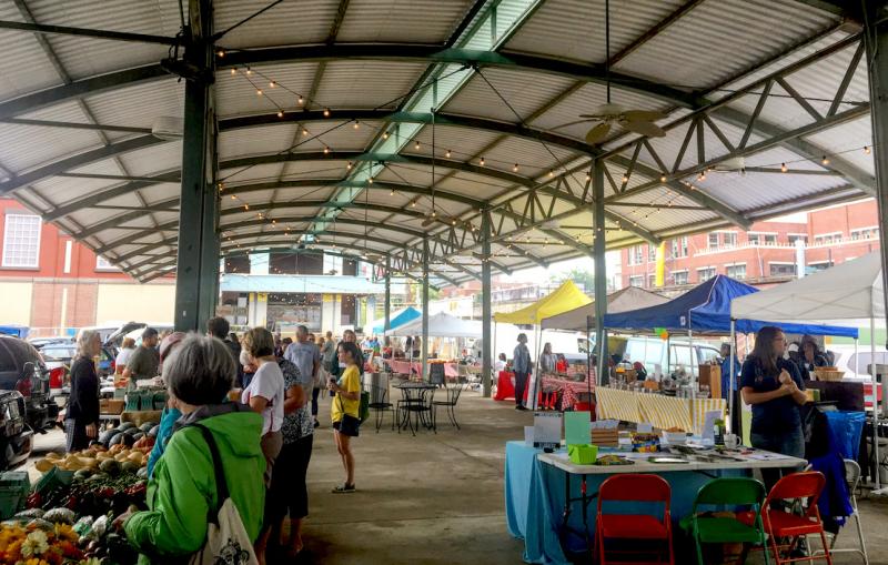 people shopping at a farmers market with booths of vegetables under an arched pavilion