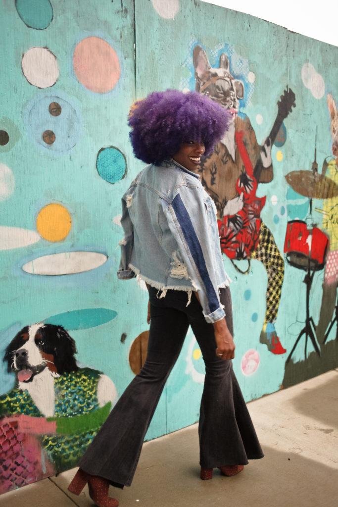 woman walking by a mural with flare-leg pants, a distressed light demin jacket, and purple afro hairstyle