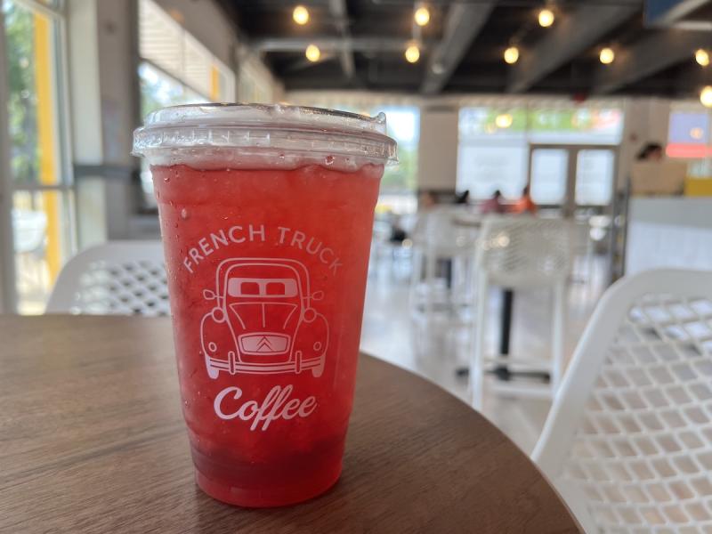 dark pink colored iced tea in a plastic cup in the foreground, background is coffee shop interior with white chairs and lots of windows