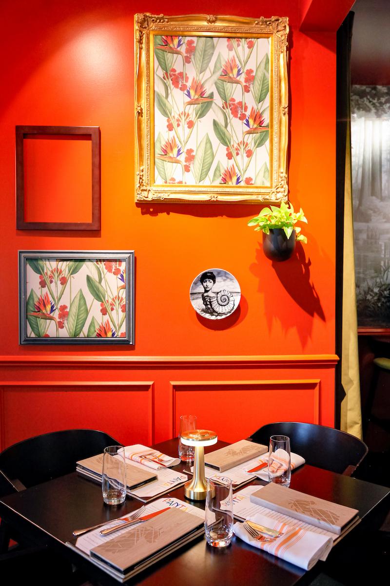 bright red-orange wall with colorful floral artwork, foreground is table set with menus