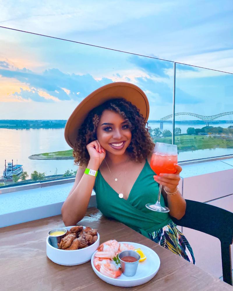 woman with brim hat dining on rooftop of building with drinks and food