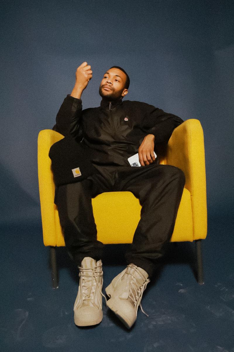 Photo of Curator Kylon Wagner. He is wearing all black with tan shoes shitting in a yellow chair