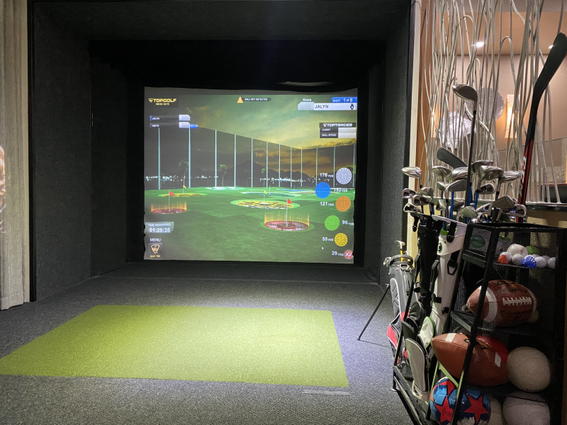 a photo of the topgolf swing suite simulator. The screen features a target golf course. Golf clubs, footballs, soccer ball, hockey puck and more athletic gear sit on a shelf
