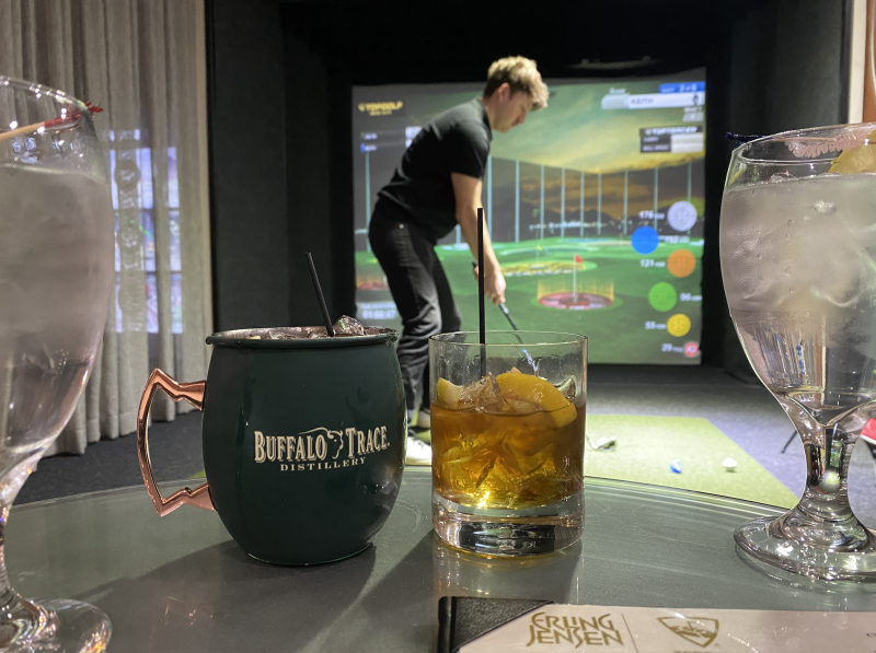 two cocktails sit on a table in front of a man playing golf on a simulator