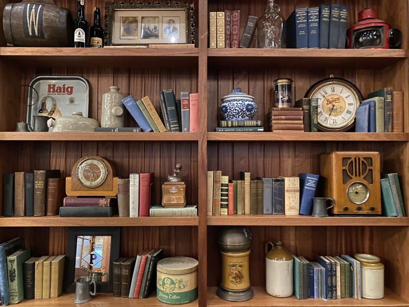 close-up photo of the bookshelf lining back wall complete with vintage looking books and knick knacks like old clocks and containers