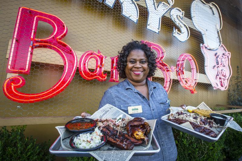 woman holds up two plates of bbq outside In front of corks sign