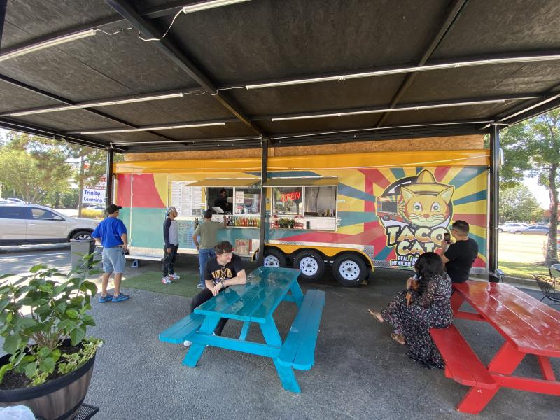 wide shot of taco cat food truck under canopy, bright colored picnic tables, people ordering and waiting