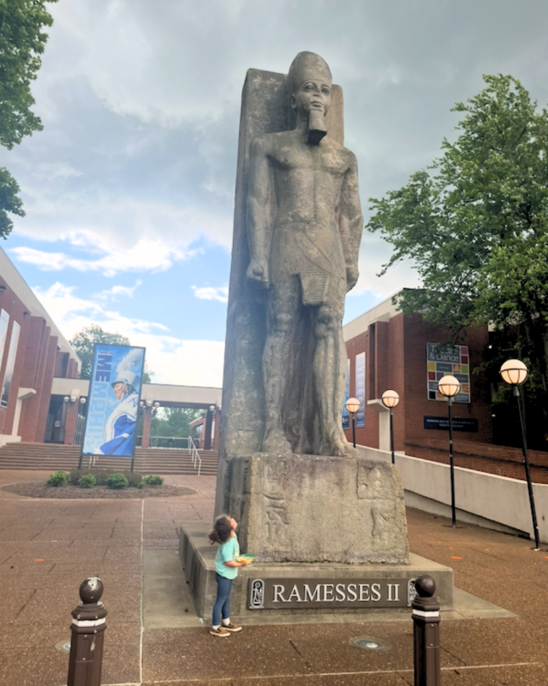 child looks up at tall  Ramesses II Statue on U of M campus
