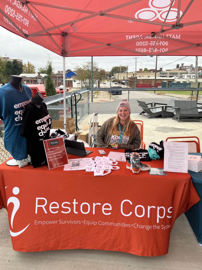 woman sits behind restore corps booth at race