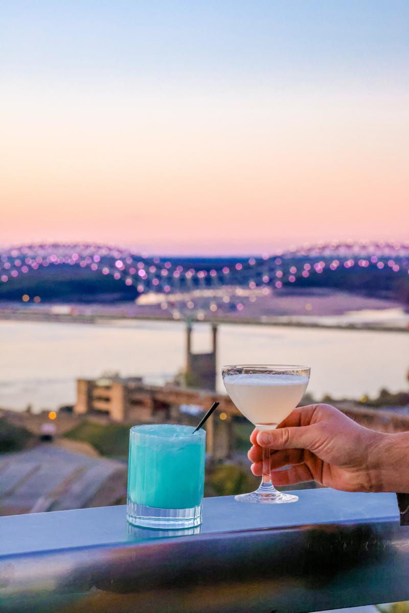 two cocktails on edge of rooftop in front of mighty lights bridge