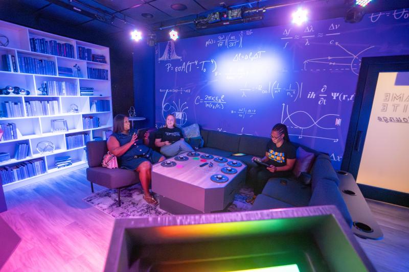 three people sit on a couch around control game panel in game nite lounge room