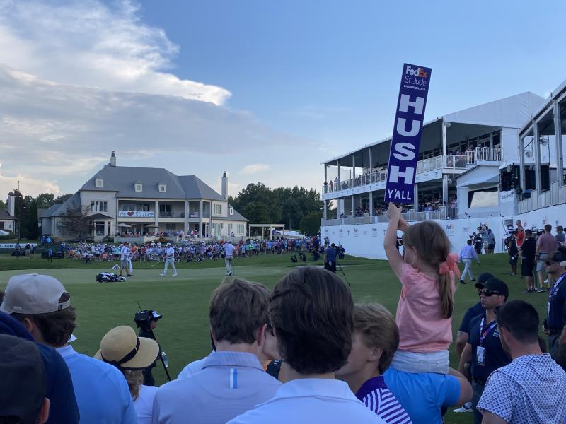 girl on dad's shoulders holds up hush y'all sign as crowd watches golfers