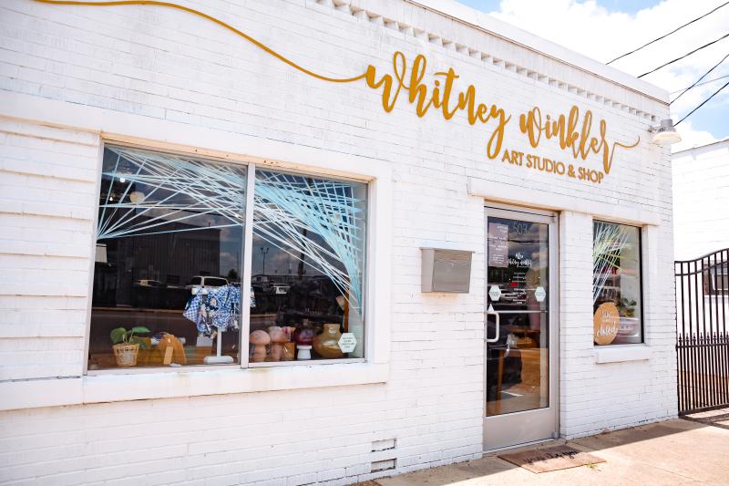 exterior of Whitney Winkler Art Studio and Gift Shop. Sign in gold script font and the building is white brick