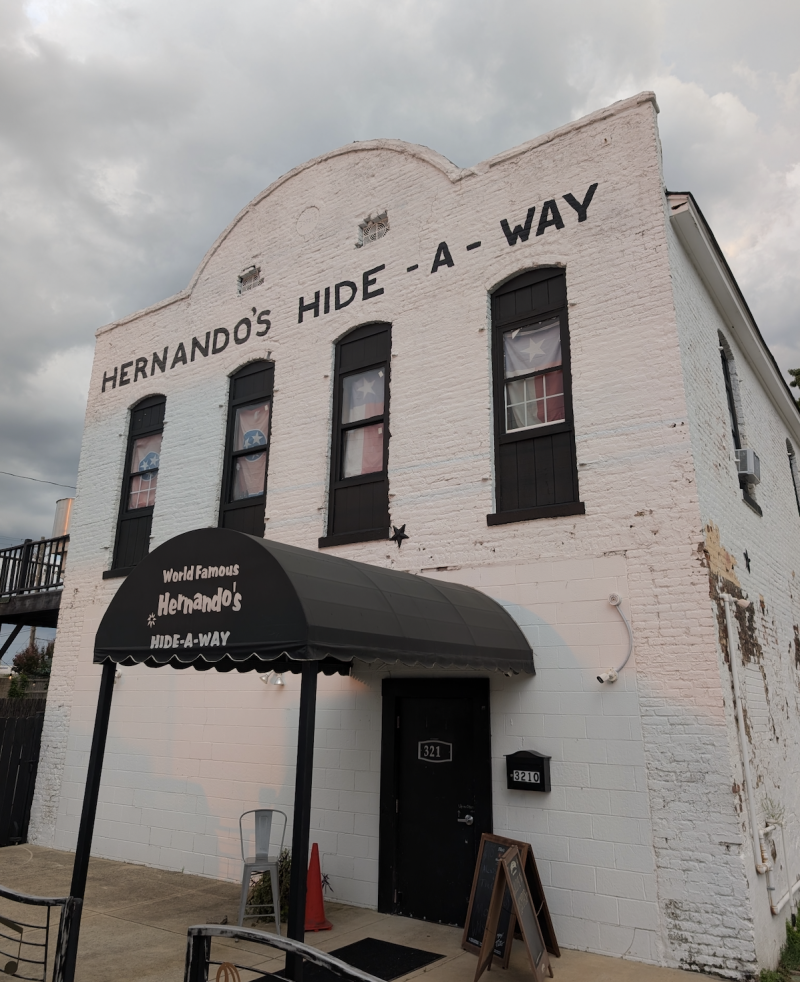 exterior of Hernando's Hide-a-way (a two story white building honky tonk with an entrance awning extending over sidewalk)