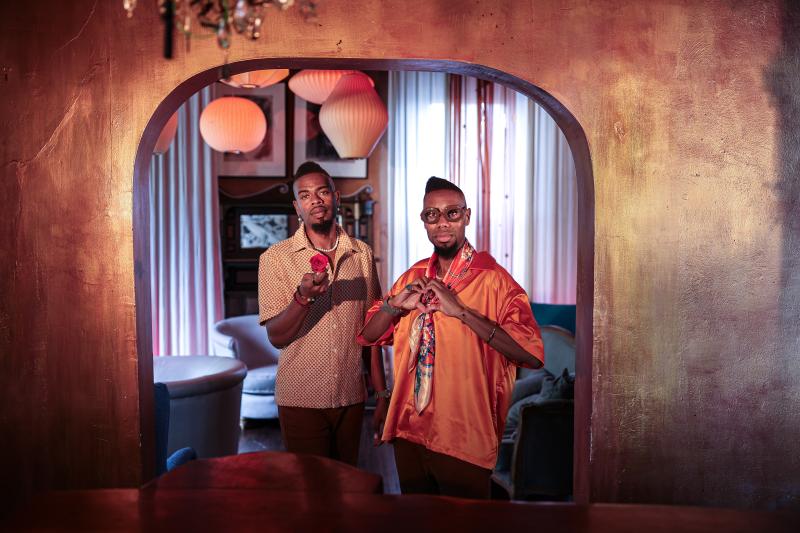two African American men standing next to each other, one making a heart symbol with his hands and one holding out a rose