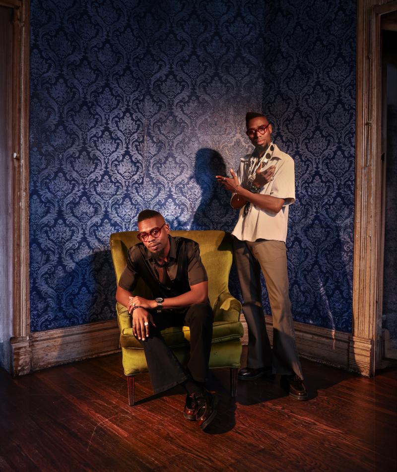 two african american men, one standing adn the other one sitting in front of blue wallpaper with a green chair