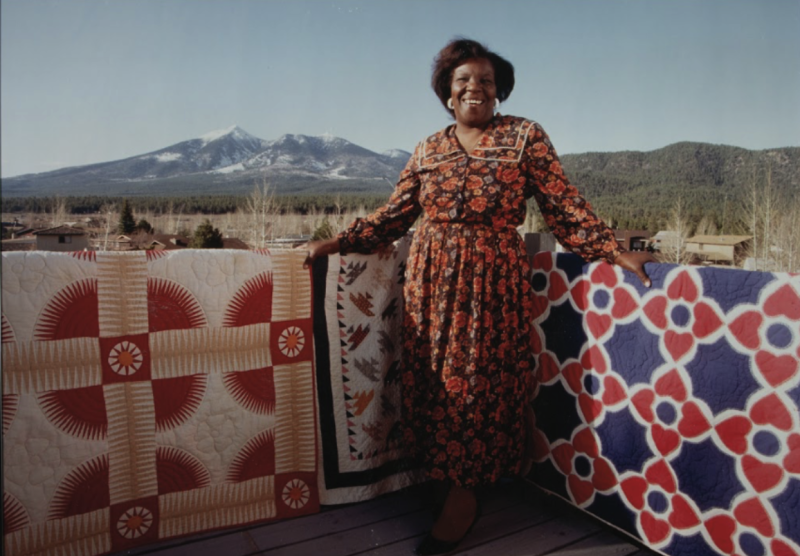 african american woman standing in front of colorful quilts with mountains in the distance behind her