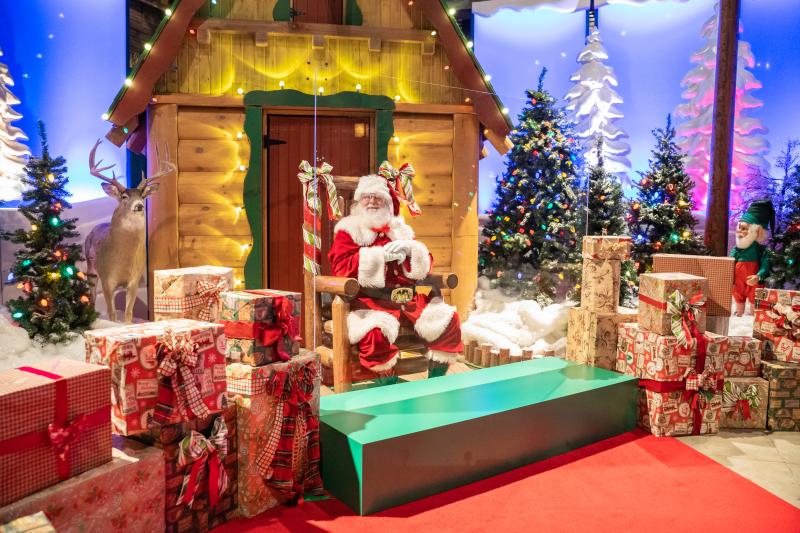 santa sitting in front of a cabin surrounded by presents
