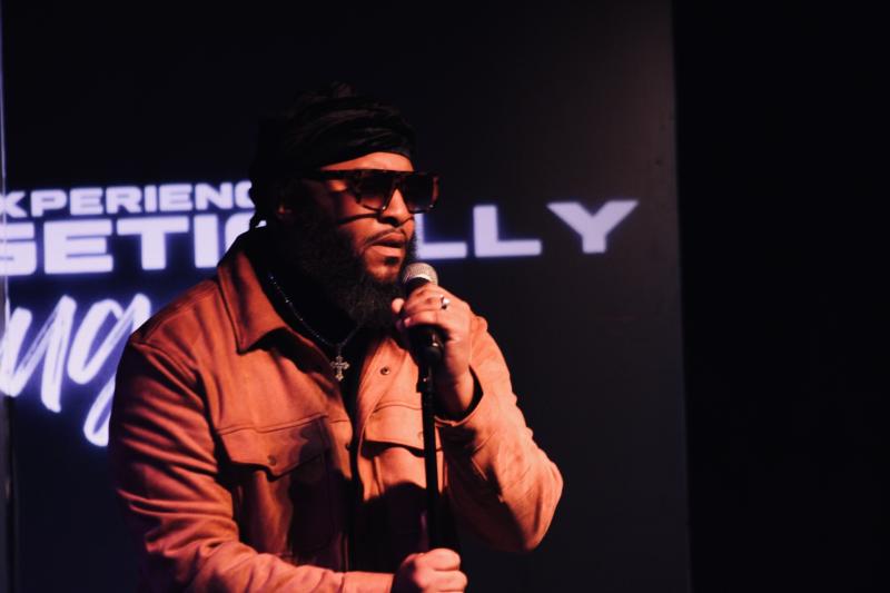 black man wearing a brown coat and sunglasses singing into a microphone