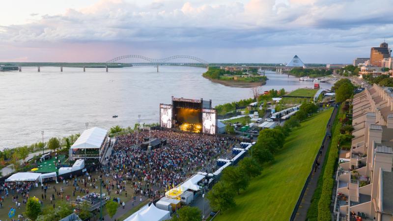 An aerial view of RiverBeat Music Fest, with a large crowd gathered in front of a large stage. The Mississippi River, Hernando de Soto Bridge and Memphis skyline are in the background.