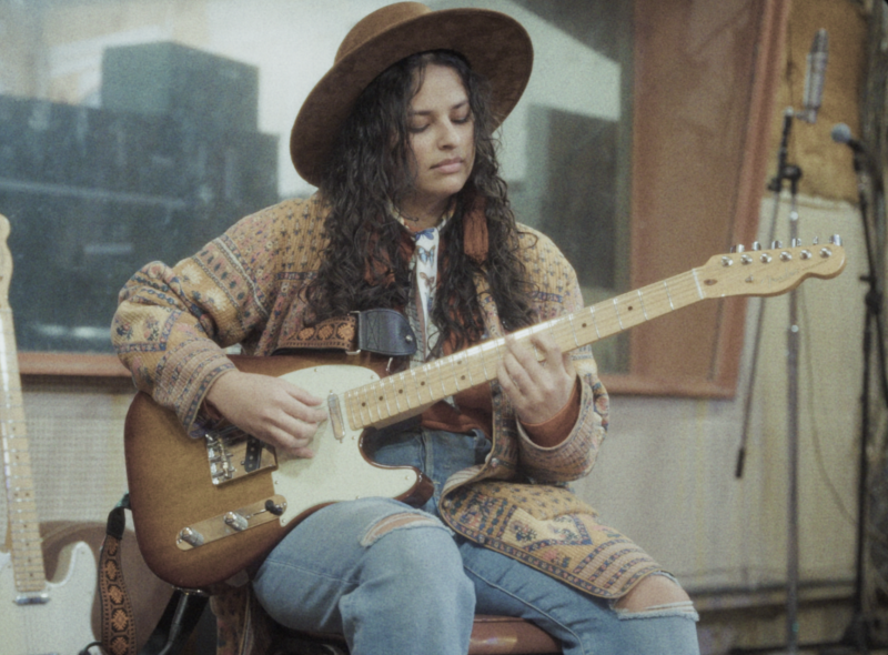 Girl with wide brimmed hat, long black curly hair, wearing oranges and browns, sitting and playing the guitar