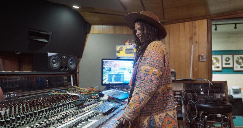 Girl with wide brimmed hat, long black curly hair, wearing oranges and browns,standing over a soundboard, looking down at all the mechanics