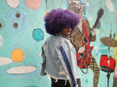 woman walking by a mural with flare-leg pants, a distressed light demin jacket, and purple afro hairstyle