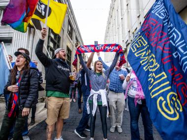 901 fc fans stand cheering in an alley during the march prior to the game. they're holding flags that say Memphis and Bluff City Mafia