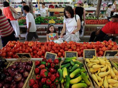 mom and daughter picking out tomatoes from large bin full. cucumbers squash red bell peppers and purple onions are in baskets in front of them