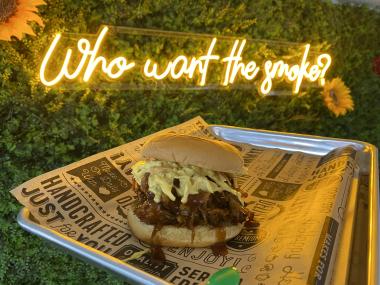 pulled pork sandwich on tray behind held up in front of "who want the smoke?" sign