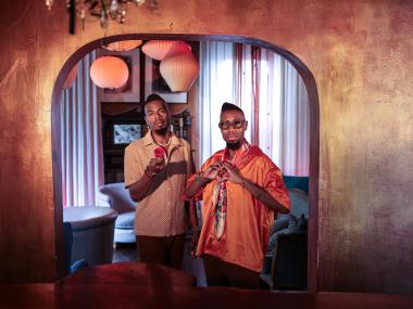 two African American men standing next to each other, one making a heart symbol with his hands and one holding out a rose