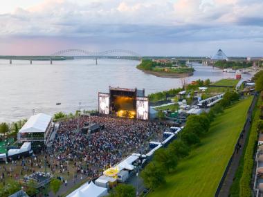 A crowd gathering at a stage at RiverBeat Music Festival at Tom Lee Park, with the Mississippi Riverfront, Downtown Memphis skyline and the Hernando de Soto bridge in the background.